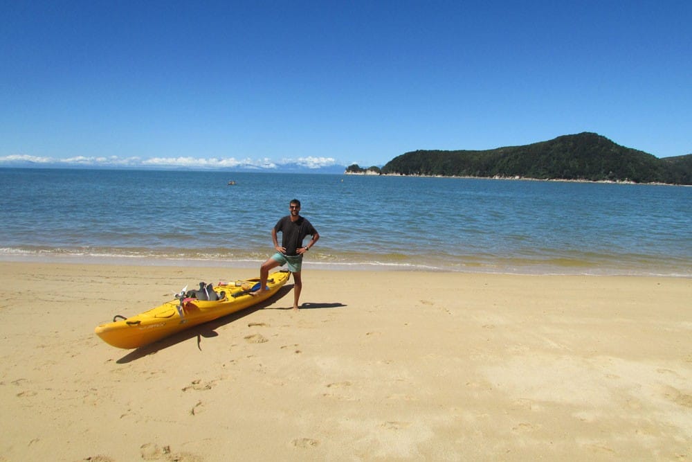Vivir en Nueva Zelanda chico moreno con un pie apoyado en un kayak y detrás la playa